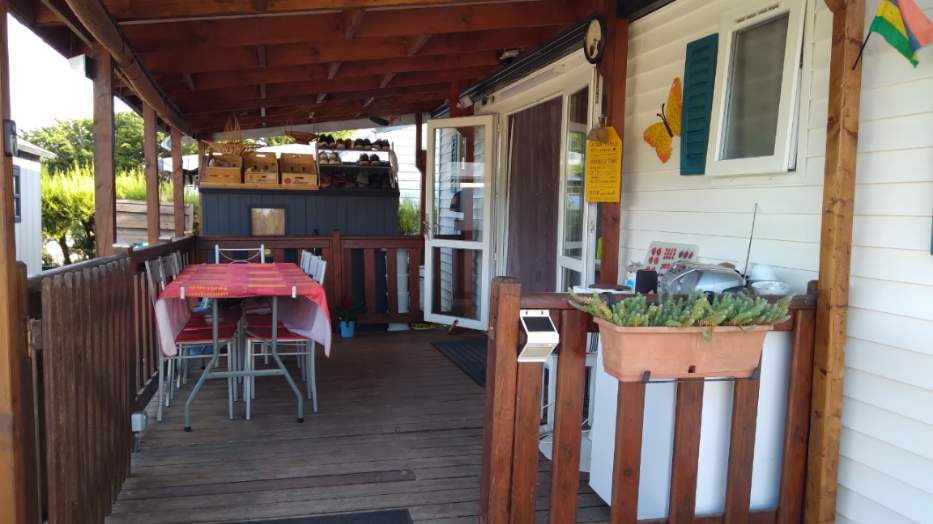 A covered terrace features a wooden deck and a table set for six with a colorful tablecloth. A planter filled with greenery is positioned on the railing, and a small sink can be seen near the wall. Light is allowed in through the glass doors.