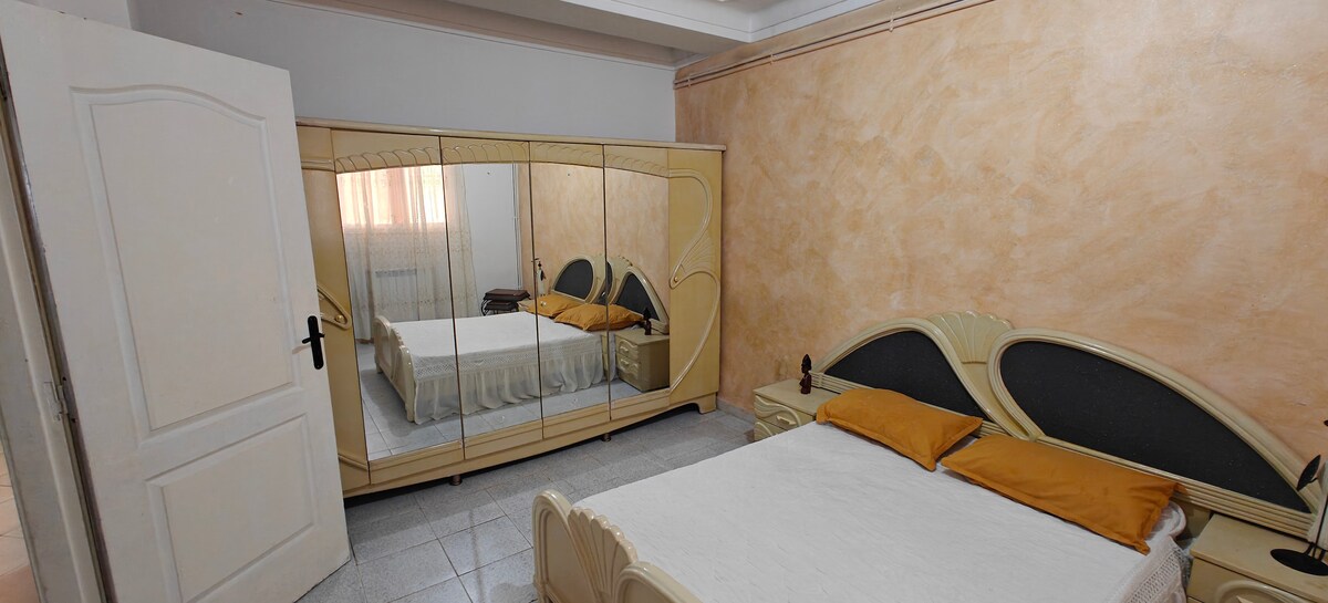 A comfortable bedroom is shown, featuring a spacious bed adorned with two yellow pillows. A large mirrored wardrobe reflects natural light from a nearby window, and light-colored walls provide a warm ambiance. The flooring is tiled, contributing to a clean and tidy environment.