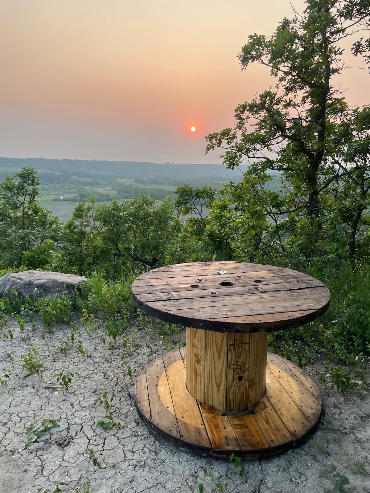 A circular wooden table made from a large spool is positioned on uneven ground, surrounded by lush greenery. In the background, a hazy sunset casts a warm glow over the distant valleys, with the natural landscape providing a serene backdrop.