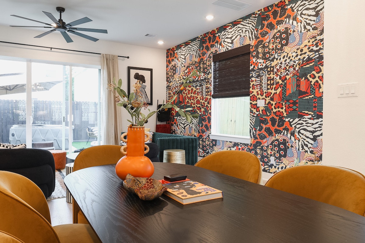 A dining area is presented with a dark wooden table and warm-toned chairs. A vibrant wall features a bold, patterned design. Natural light enters through a large window, with a view of the backyard visible. A decorative vase with greenery sits at the center of the table.