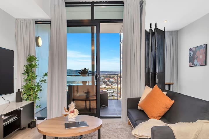 Svetlá, moderná obývačka s bielou koženou pohovkou a dreveným konferenčným stolíkom. Posuvné sklenené dvere sa otvárajú na súkromný balkón s výhľadom na mesto a napĺňajú priestor prirodzeným svetlom. Ideálne na relaxáciu alebo zábavu v štýlovom a pohodlnom prostredí.