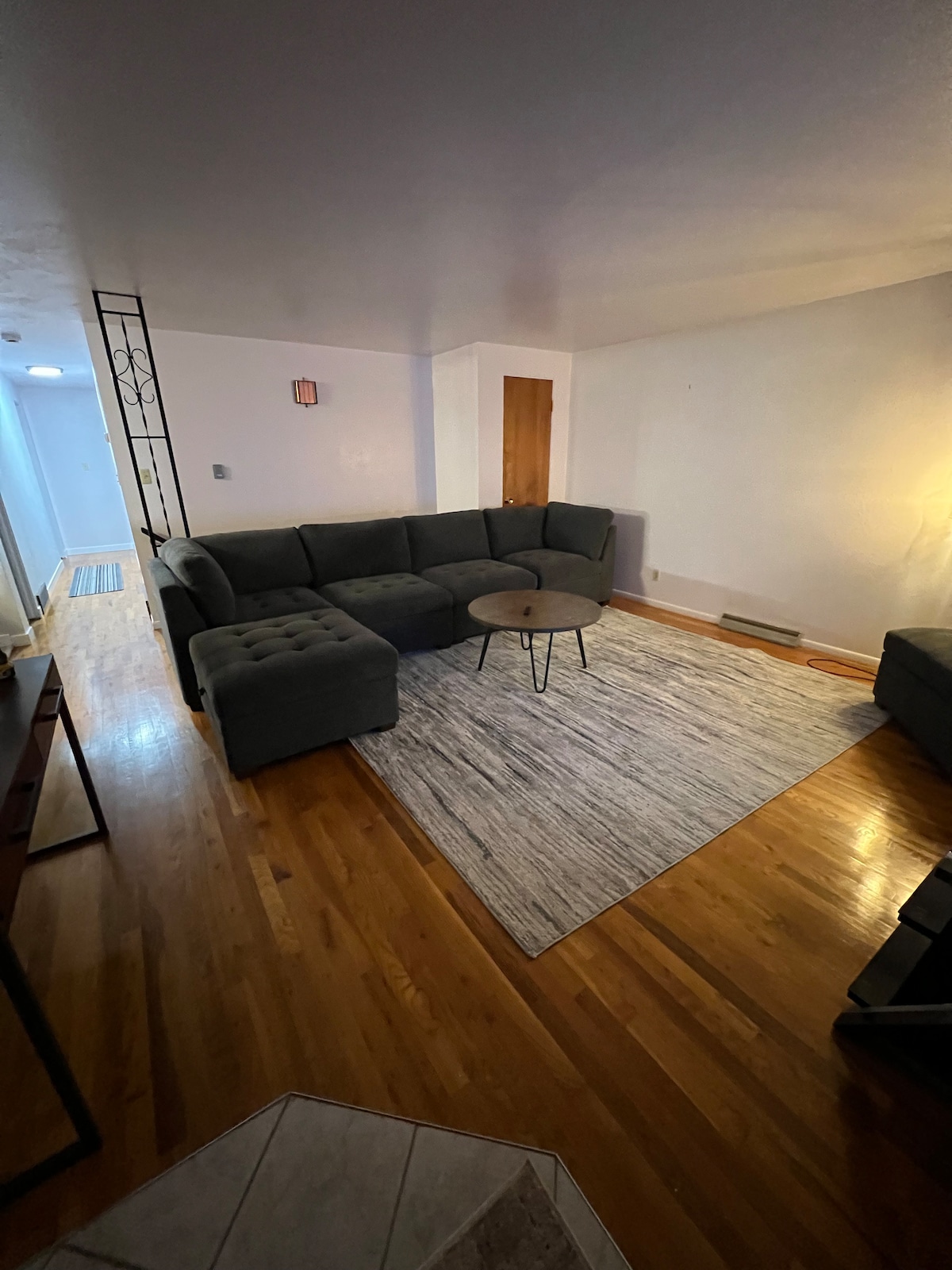 A comfortable living area with a sectional sofa and a round coffee table is present. The hardwood flooring is accentuated by a light-colored area rug. A warm light source from a nearby lamp gently illuminates the space, creating a welcoming environment.