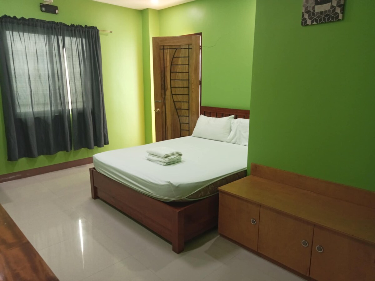 A sleeping area features bold green walls complemented by a spacious bed adorned with tidy white bedding. A wooden dresser and a set of shelves provide practical storage solutions. Natural light filters through a window dressed with dark curtains.