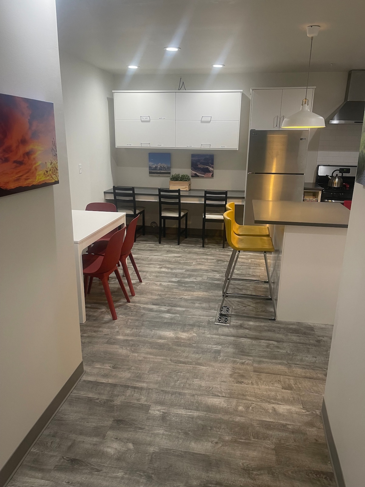 A modern kitchen space is displayed, featuring a table with mixed seating of red and black chairs. Yellow bar stools are positioned at the counter. The sleek cabinetry above and stainless steel appliances create a functional yet inviting atmosphere, with light streaming from overhead fixtures.