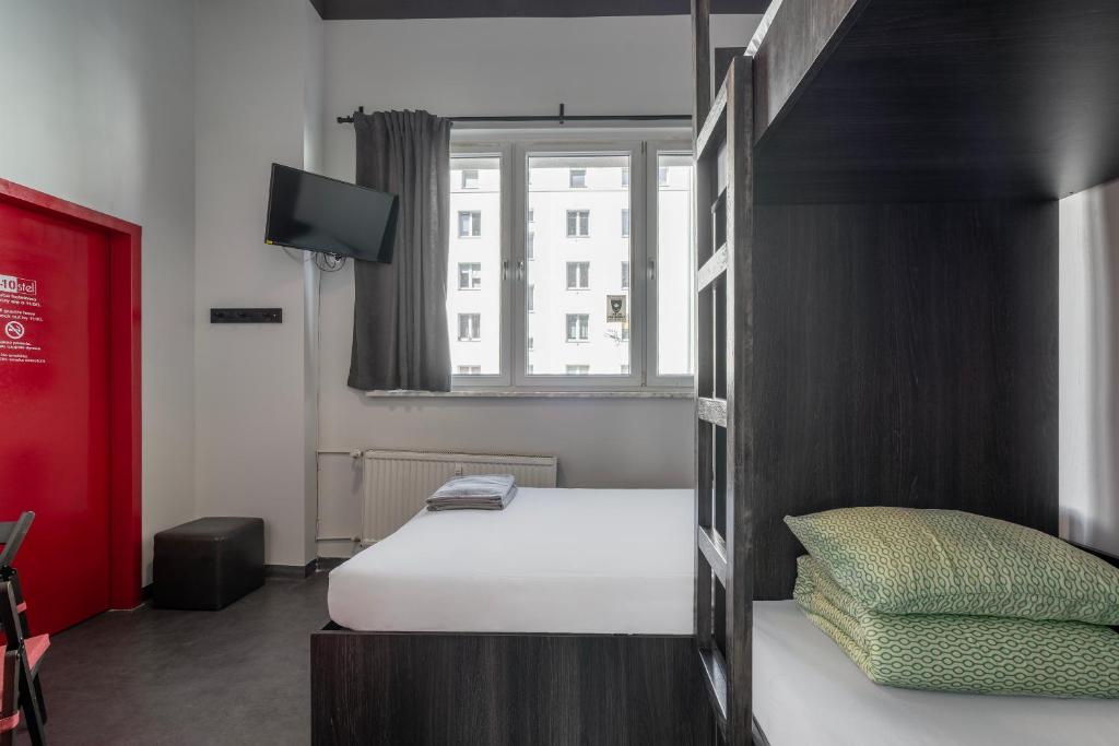 A minimalist guest room features a spacious bed with neatly arranged green-patterned pillows. A wall-mounted television is visible above a black wooden closet. Natural light enters through a large window dressed with gray curtains, illuminating the room's clean and modern design.