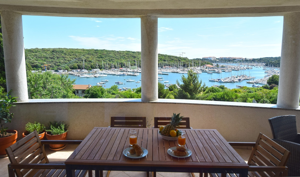 A spacious balcony is presented with a wooden dining table, featuring two glasses of orange juice and pastries. Lush greenery and a marina filled with boats are visible in the background, with gentle hills framing the picturesque view under a clear blue sky.
