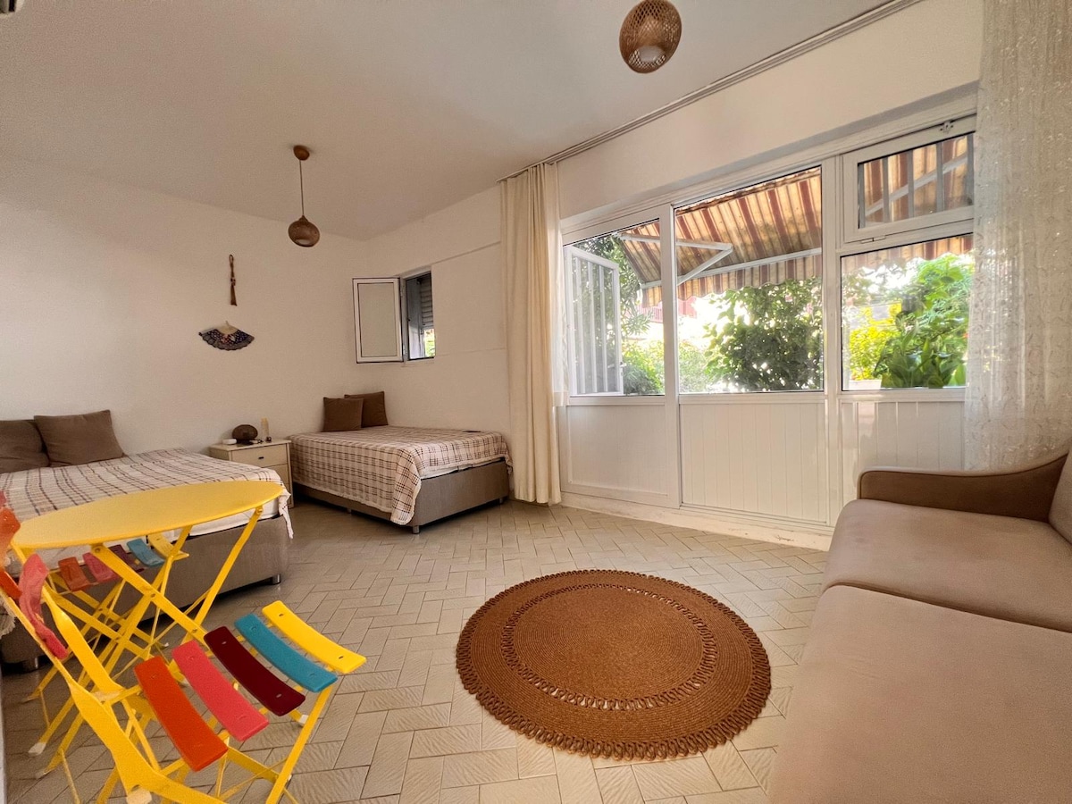 A spacious room features two beds with neutral-toned bedding and a comfortable sofa. A colorful folding table and chairs are arranged nearby. Large windows allow natural light, offering views of greenery outside. Textured flooring and a circular rug add warmth to the space.