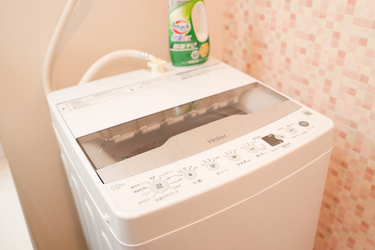 A washing machine is situated within the accommodation. The machine features a clear lid allowing visibility of the interior, along with various settings displayed on the front panel. A bottle of detergent is placed on top for convenient access.
