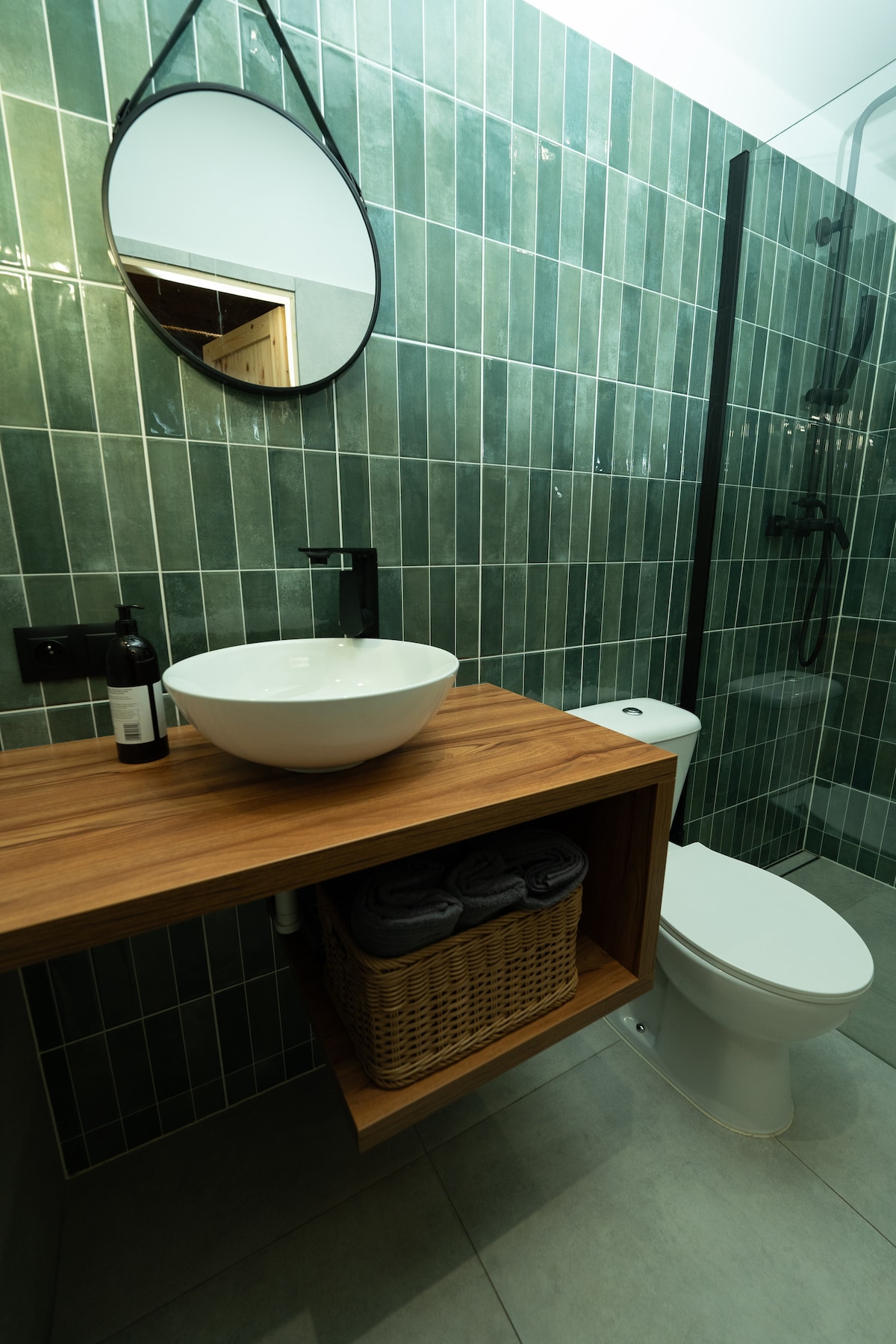 A modern bathroom features green tiled walls and a sleek, wooden vanity. A round mirror is mounted above a white basin sink, while a shower area is enclosed with glass. A basket holds neatly stacked towels next to the toilet, enhancing the organized space.
