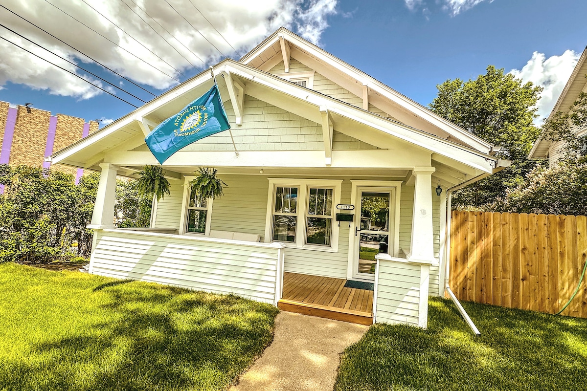 A charming historic bungalow presents a welcoming front porch with hanging plants. A bright blue sky with scattered clouds enhances the home's fresh appearance. A large flag is displayed prominently, and a wooden fence encloses the yard, which features well-maintained green grass.