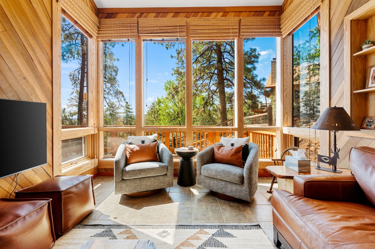 A bright sunroom features large windows surrounded by rustic wood paneling, allowing natural light to fill the space. Comfortable chairs are positioned around a small table, creating an inviting area for relaxation. A cozy leather sofa and a decorative rug complement the serene atmosphere.