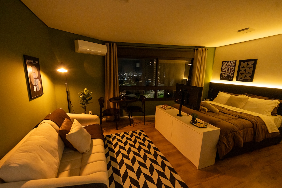 A modern studio space features a spacious bed with neutral bedding against a green accent wall. A cozy seating area is highlighted by a sofa and a small table, creating an inviting atmosphere. Large windows provide views of the city skyline, complemented by soft lighting.