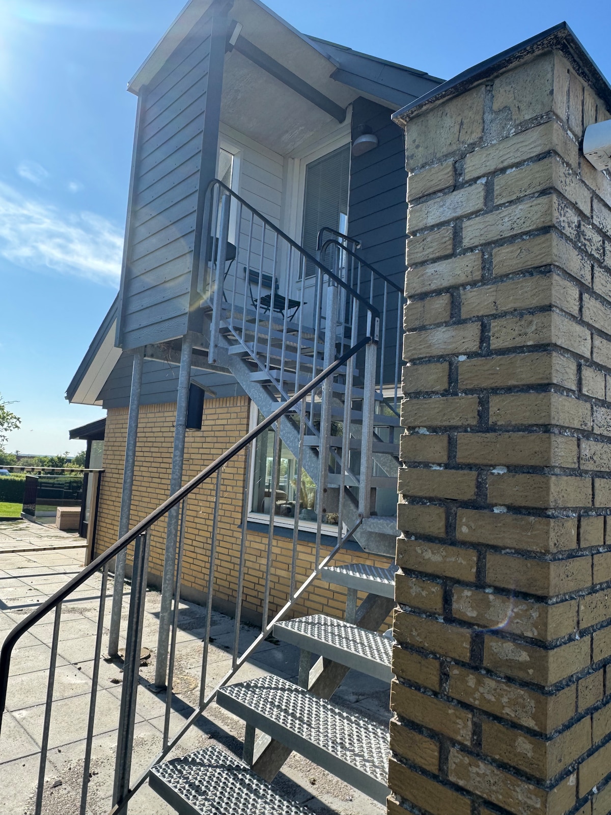 An exterior view of the building features a set of metal stairs leading to an upper entrance. The staircase is bordered by a brick wall, and large windows are visible on the upper floor, allowing ample natural light to brighten the space.