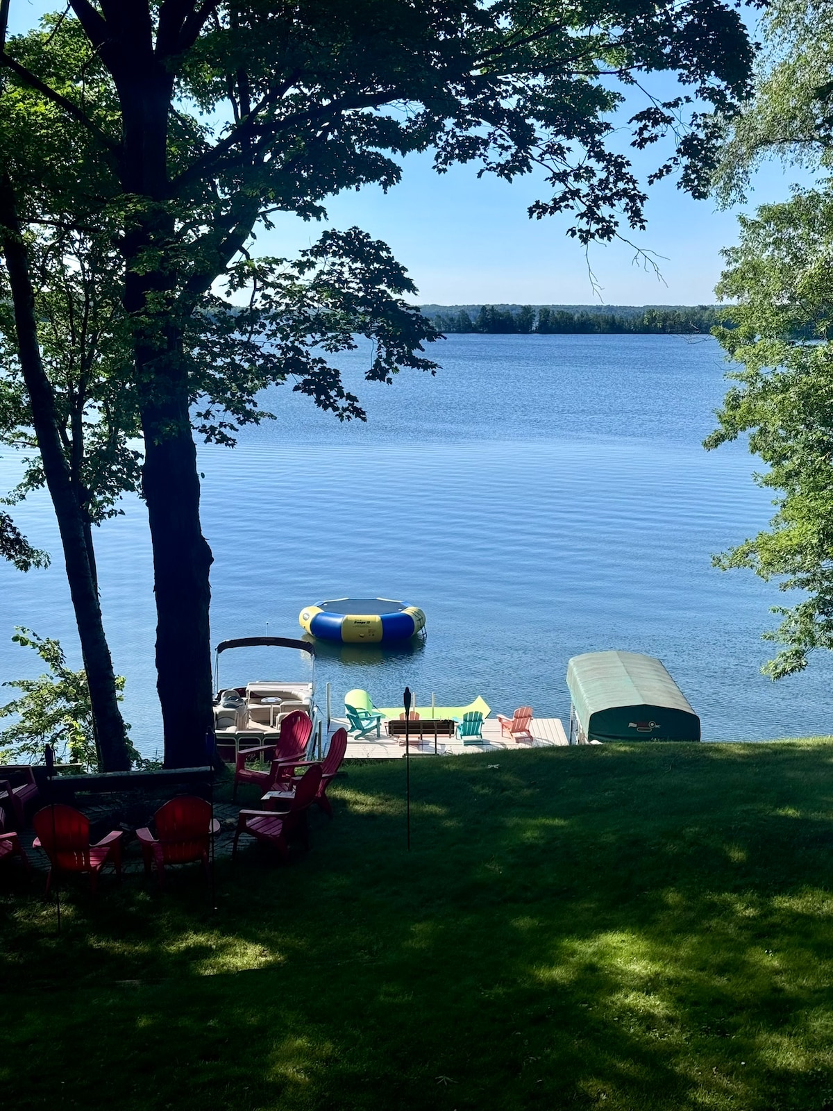 A serene lakefront scene is presented, featuring calm waters reflecting the blue sky. A swim raft floats on the water near a shaded dock, with vibrant green grass and a gathering of colorful chairs arranged around a fire pit. Trees provide natural framing.