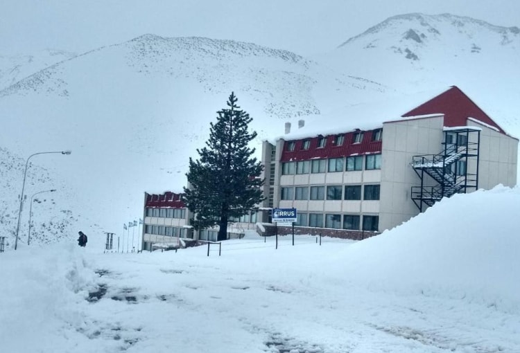 A snow-covered exterior of the Cirrus building is visible, set against a backdrop of snow-capped mountains. The structure displays a red-roofed facade, with a set of stairs accessible from the ground level. The pathway is outlined by freshly fallen snow, enhancing the serene winter landscape.