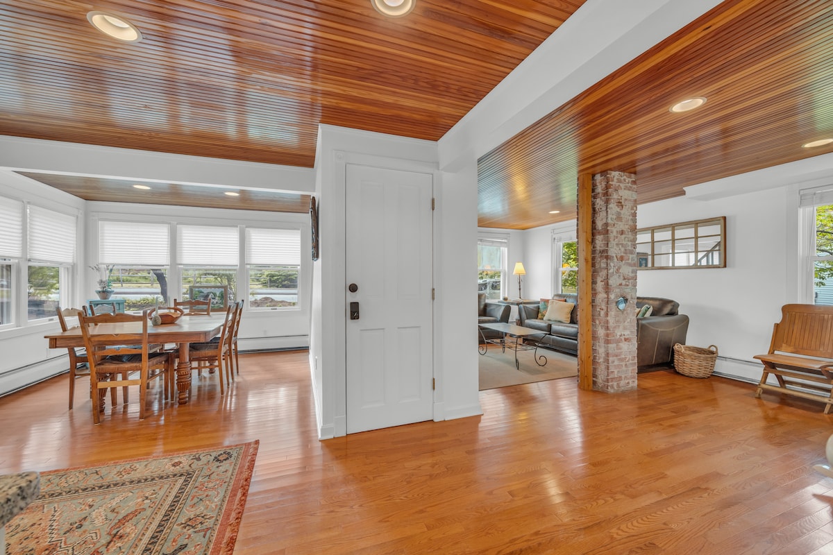 The open living space features hardwood flooring and wood-paneled ceilings. A dining area with a wooden table and chairs is beside large windows allowing natural light. A living area is visible in the background, with a comfortable sofa and a brick column adding character.