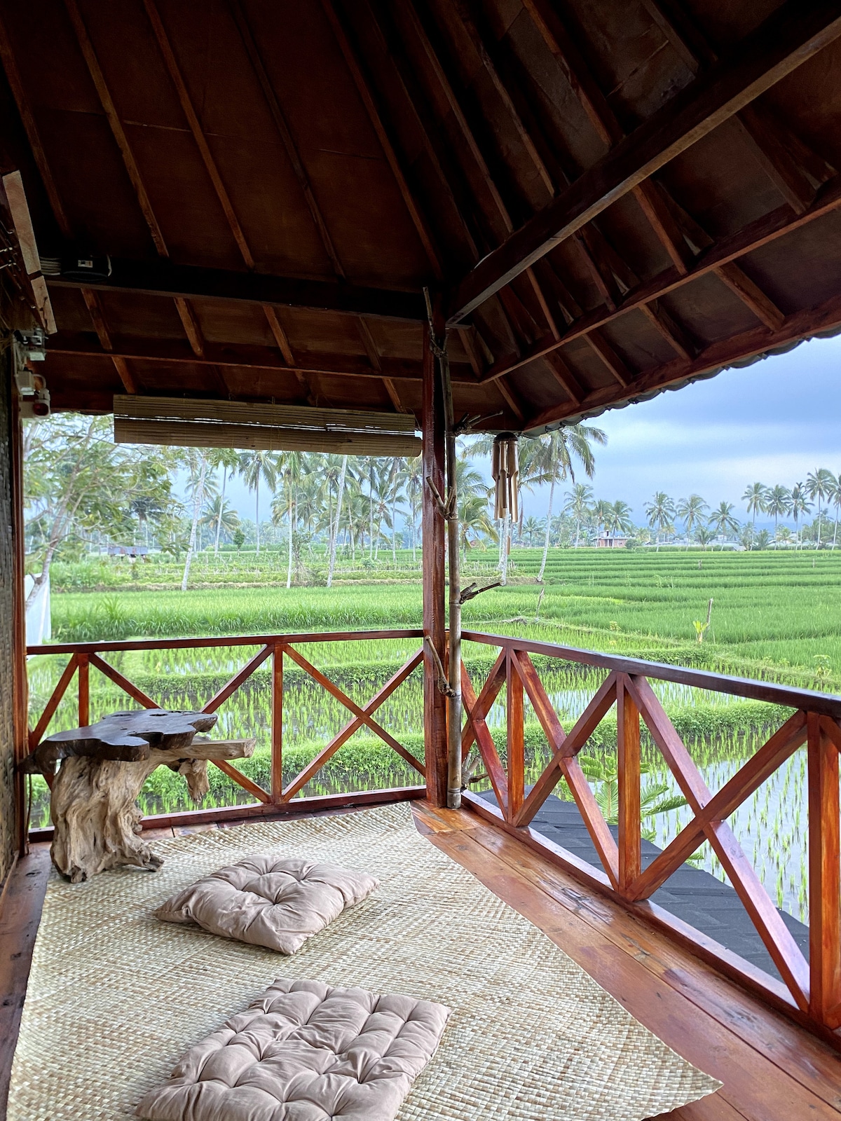 A cozy balcony is presented with traditional wooden beams and a thatched roof, offering views of lush rice fields. Cushioned seating is arranged on a bamboo mat, inviting relaxation. Green palm trees dot the landscape, contributing to a serene atmosphere.