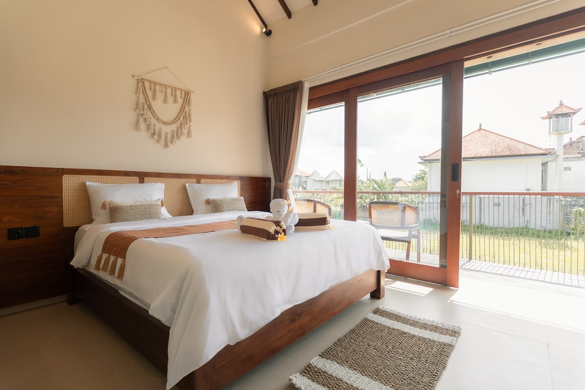 A spacious bedroom features a large bed with neutral bedding and decorative accents. Floor-to-ceiling glass doors open to a balcony overlooking rice fields, allowing natural light to fill the room. A woven rug lies on the tiled floor, complementing the warm wood furnishings.