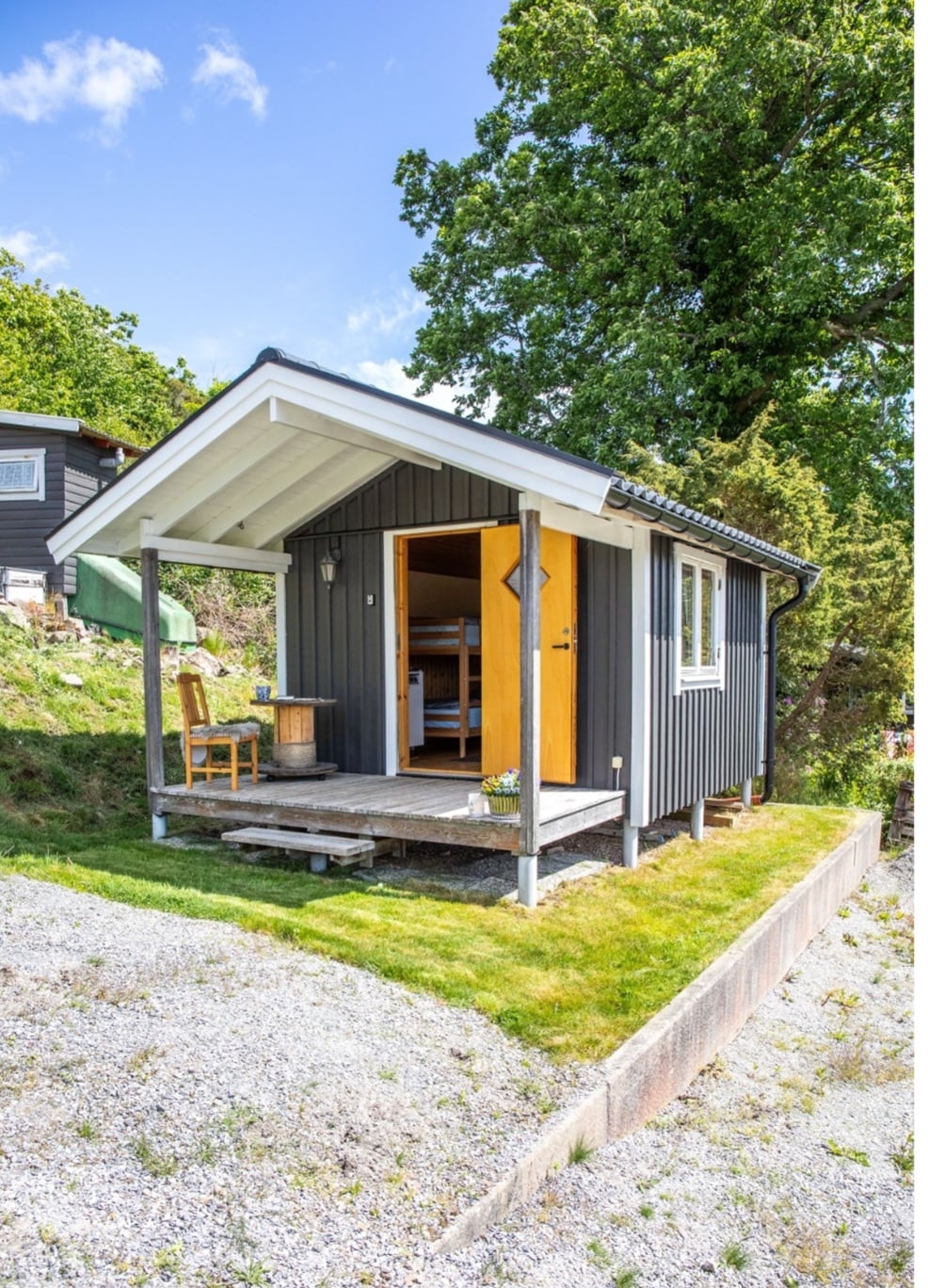 A compact guest cabin is shown with a small porch featuring a wooden chair. The cabin's door is open, revealing the interior. Surrounding greenery complements the cabin's dark wood exterior, and a well-maintained gravel pathway leads to the entrance.