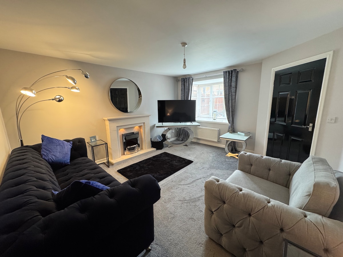 A living room features a modern, cozy atmosphere with a black sectional sofa and a light gray armchair. A decorative lamp stands near the fireplace, which is complemented by a round mirror above it. A television is mounted on the wall, providing entertainment options for guests.