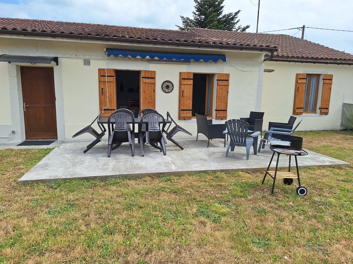 An outdoor patio area features a spacious concrete surface with several chairs and tables arranged for relaxation. Two lounge chairs and a barbecue grill are positioned nearby. The house displays wooden shutters on its windows, adding a rustic charm.