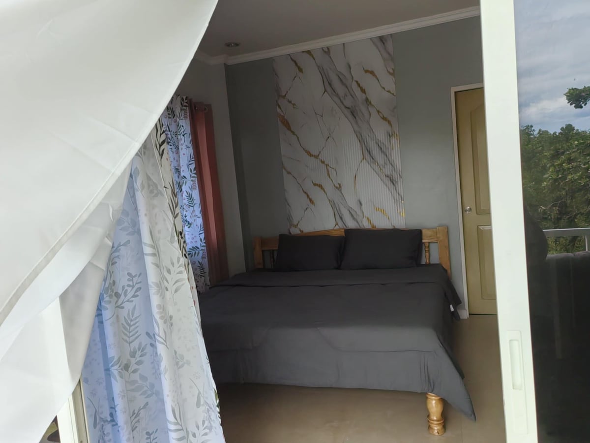 A spacious sleeping area features a large, dark-toned bed with coordinated bedding. The room displays a marble-patterned accent wall and natural light flows through the partially drawn curtains, creating a serene environment. A door leads to an outdoor view.