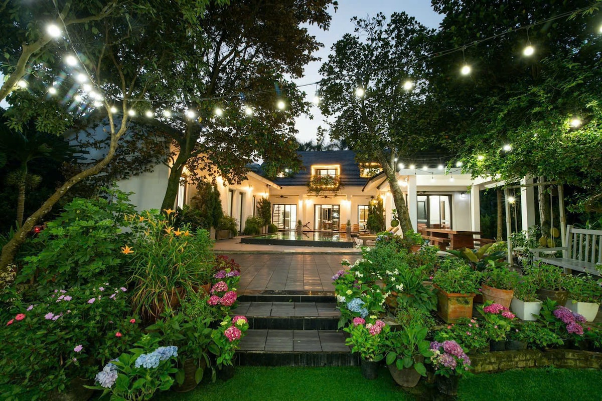 A garden space is framed by lush greenery and colorful flowers leading to a welcoming entrance of the villa. String lights are draped overhead, adding illumination to the serene setting as twilight approaches. The patio presents an inviting area for gatherings and relaxation.