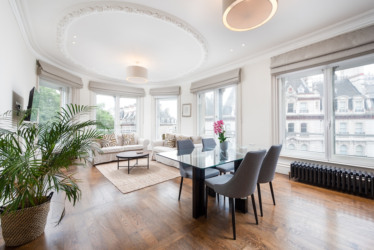 A modern living area features high ceilings with elegant crown molding and floor-to-ceiling windows providing ample natural light. A dining table with six chairs is positioned adjacent to a comfortable seating area, which includes a pair of patterned sofas. A plant adds a touch of greenery.