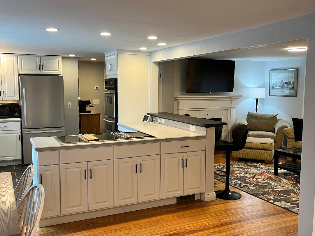 The kitchen area features white cabinetry and a spacious countertop, equipped with modern appliances and a stovetop. An open layout leads to a cozy living space, visible in the background, with comfortable seating and a mounted television above a fireplace.