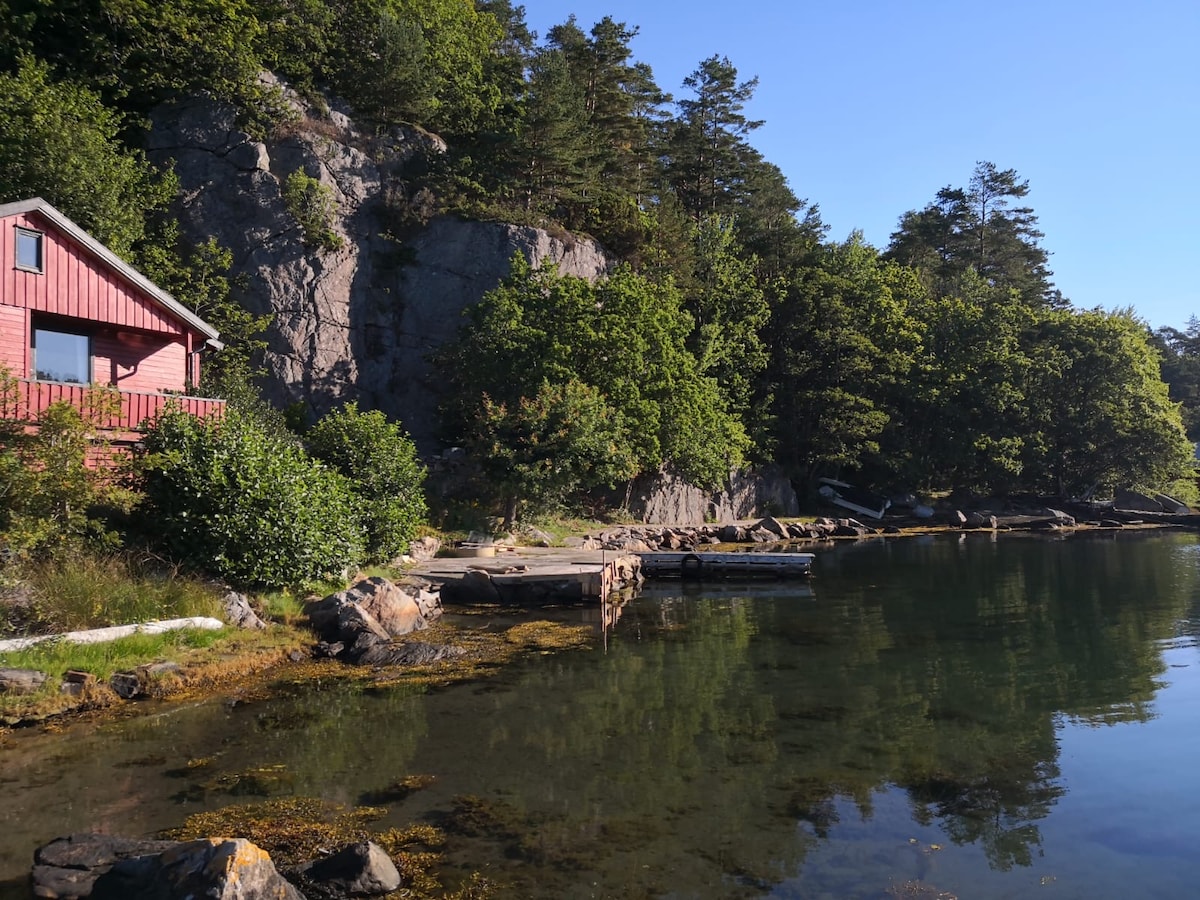 A red cabin is nestled near a rocky cliff, surrounded by lush greenery. The calm water reflects the natural landscape, with wooden docks extending into the water. Sunlight casts a warm glow over the serene scene, creating a peaceful coastal atmosphere.