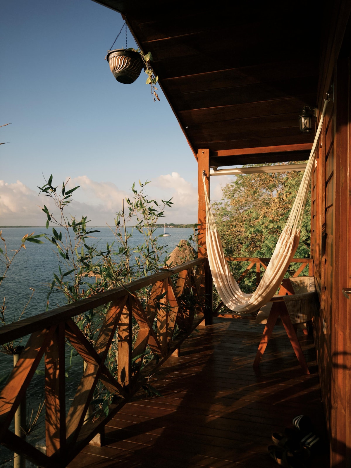A wooden balcony offers scenic views of the water, complemented by a hanging hammock and a small table. Lush greenery surrounds the space, creating a serene atmosphere. Natural light enhances the warm tones of the wooden railing and flooring, inviting relaxation.