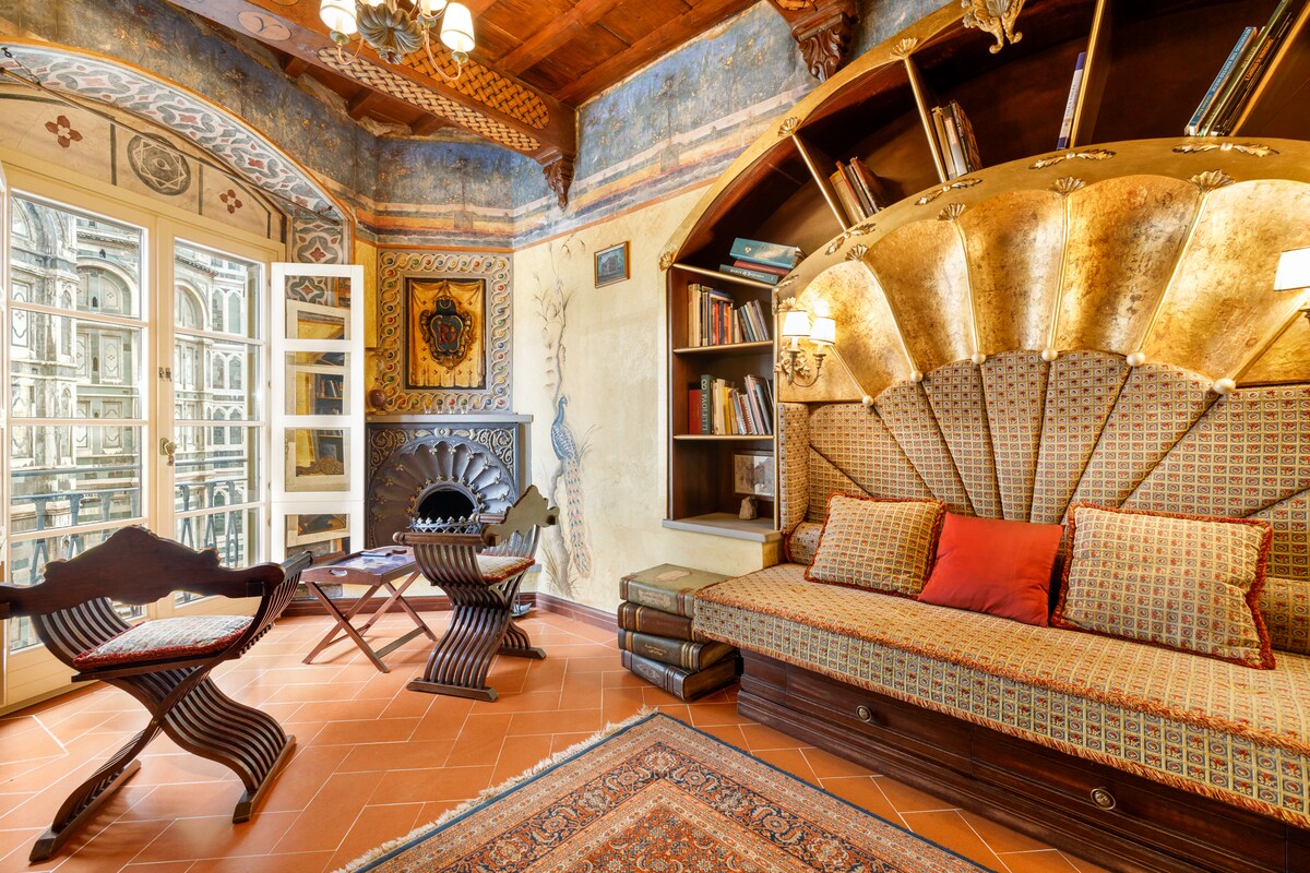 The sitting area features a shell-shaped sofa with intricate gilded details and patterned upholstery. Two wooden chairs are positioned nearby, complementing a small table. A black fireplace with decorative motifs is visible, and a view of the outside is framed by french windows adorned with elegant shutters.