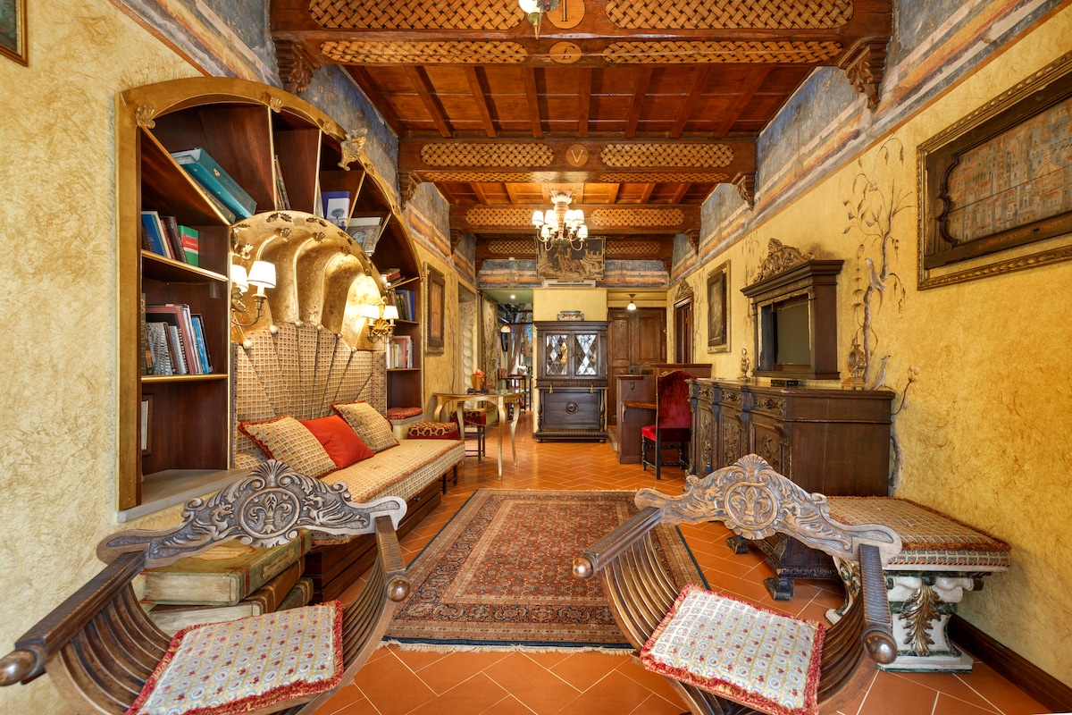 A sitting room features ornate wooden beams on the ceiling and rich golden walls. A shell-shaped sofa is positioned prominently, surrounded by decorative wooden furnishings and a bookshelf. A classic rug covers the floor, adding warmth. Wooden accents infuse the space with historical charm.
