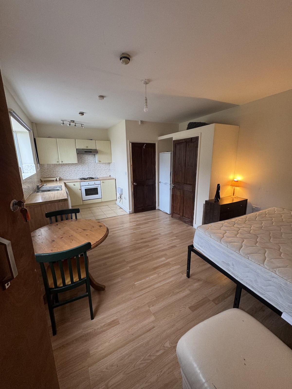 A well-lit studio apartment features a dedicated workspace, a round dining table with two green chairs, and a spacious bed with a bare mattress. The kitchen area contains fitted cabinets, a stove, and a refrigerator. Two wooden doors are visible, providing access to additional storage.