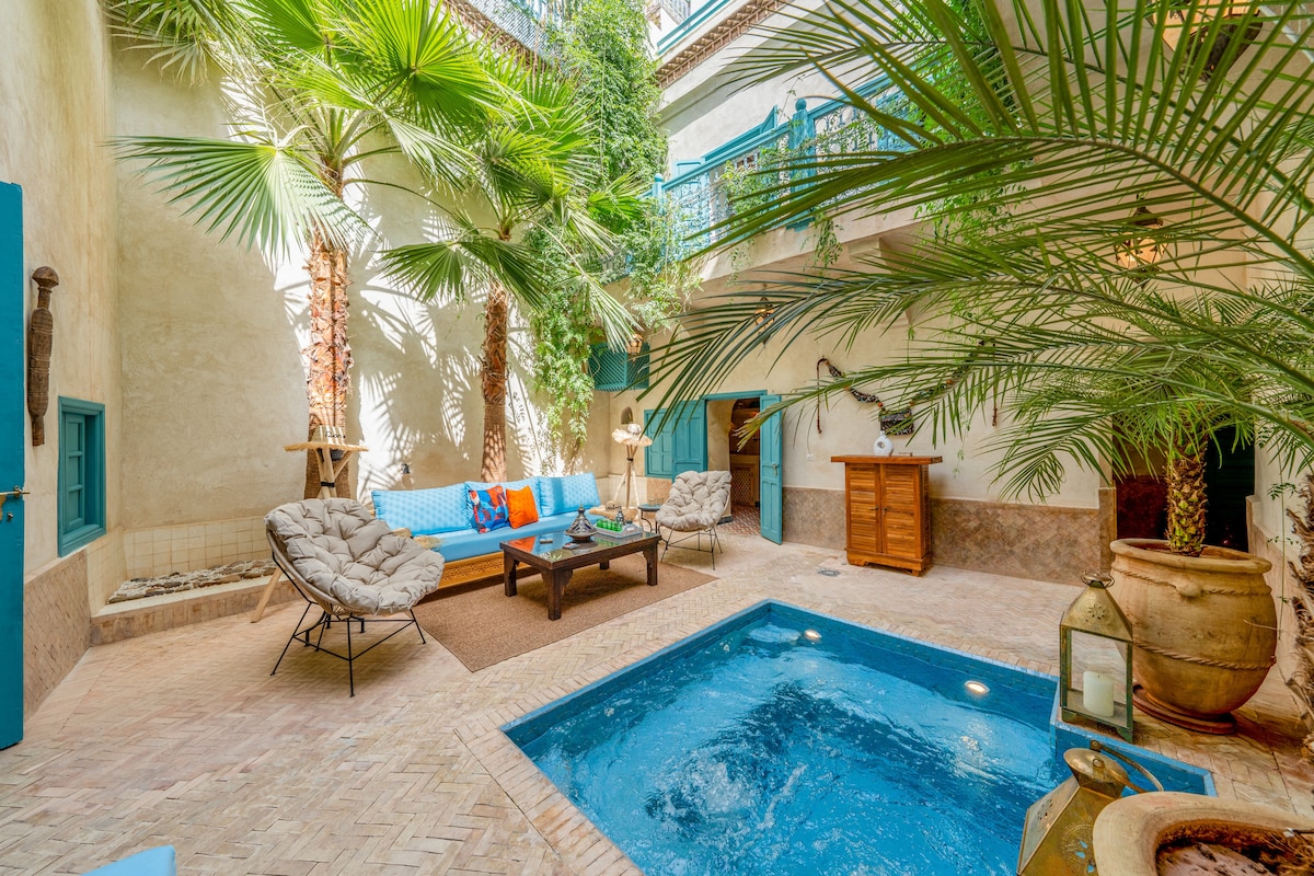 A central courtyard is surrounded by lush green palms, featuring a jacuzzi pool framed by beige tiles. A blue sofa adorned with colorful cushions offers seating alongside two comfortable chairs. Traditional décor elements are visible, enhancing the serene space filled with natural light.