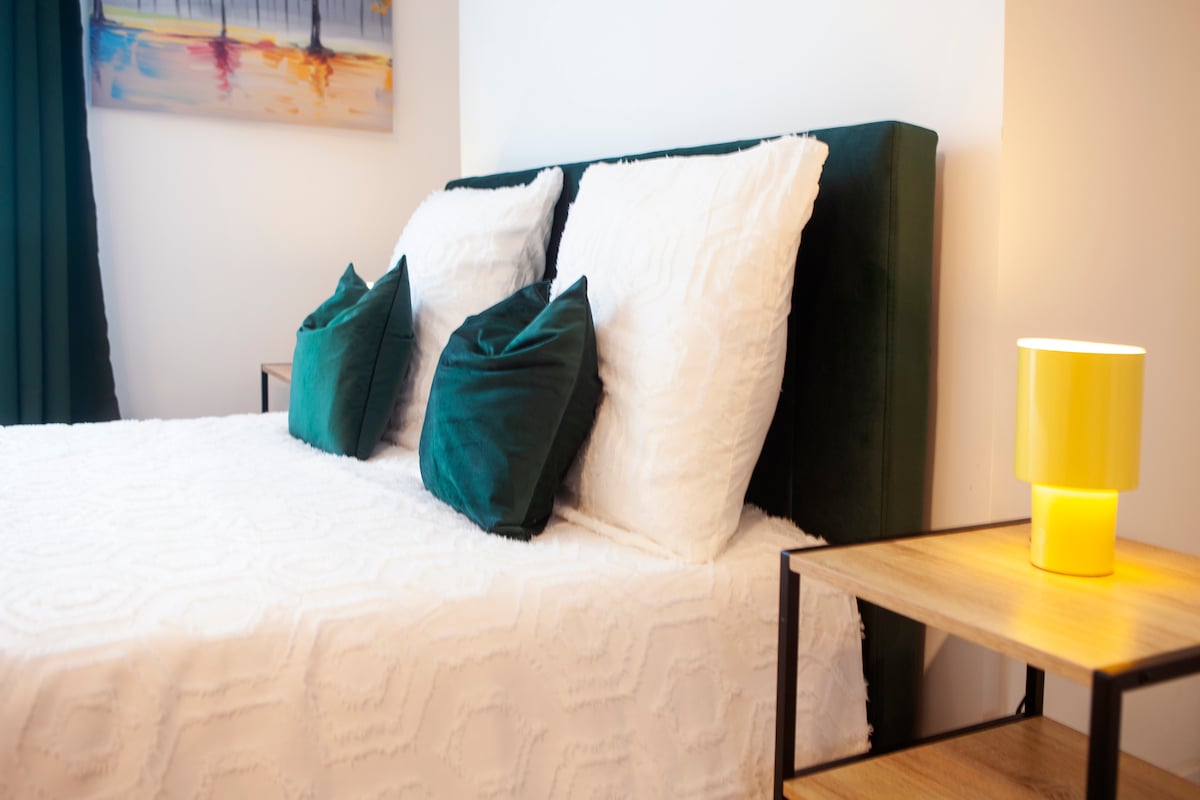 A spacious bed is dressed in white linens, complemented by several decorative cushions in deep green. A small wooden bedside table with a yellow lamp sits beside the bed, adding a touch of color to the calming atmosphere. Soft lighting enhances the inviting ambiance.