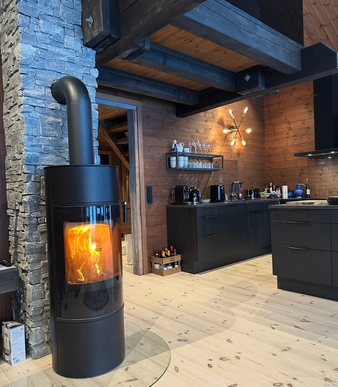 A modern black fireplace is positioned against a stone wall, providing a cozy focal point. Warm flames are visible through the glass front. A sleek kitchen area is nearby, featuring dark cabinetry and various kitchen appliances arranged on a counter.