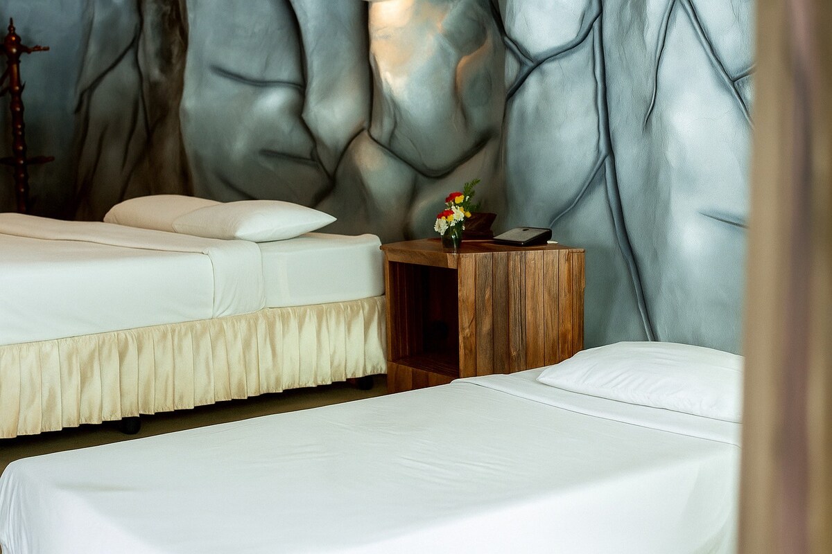 A spacious room features a king bed alongside a semi bed, both dressed in crisp white linens. A wooden nightstand holds a small flower arrangement, contributing to the inviting atmosphere. The artistic wall design adds a unique element to the overall decor.