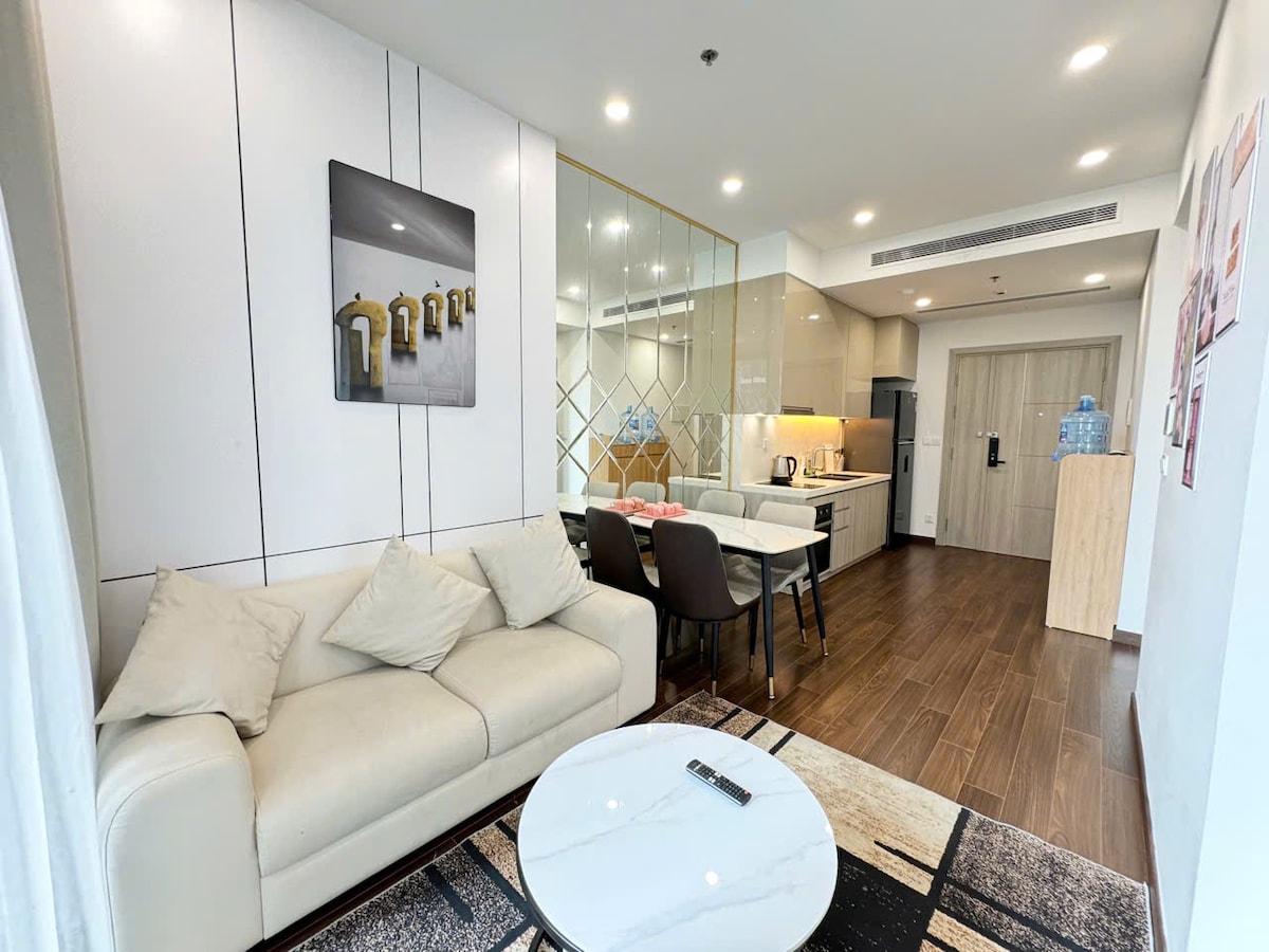 A bright living area showcases a comfortable white sofa with decorative cushions positioned near a marble coffee table. Reflections from glass accents create an open feel, while a dining area is visible in the background. Modern kitchen appliances are neatly organized, contributing to the sleek design.