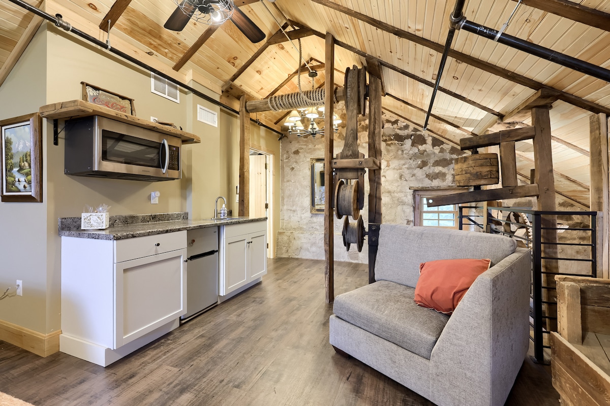 An open loft space features a kitchenette with a microwave and sink, complemented by a stone wall that adds character. A cozy gray sofa with a vibrant accent pillow is positioned near a wooden structure, creating a blend of rustic and modern design elements.