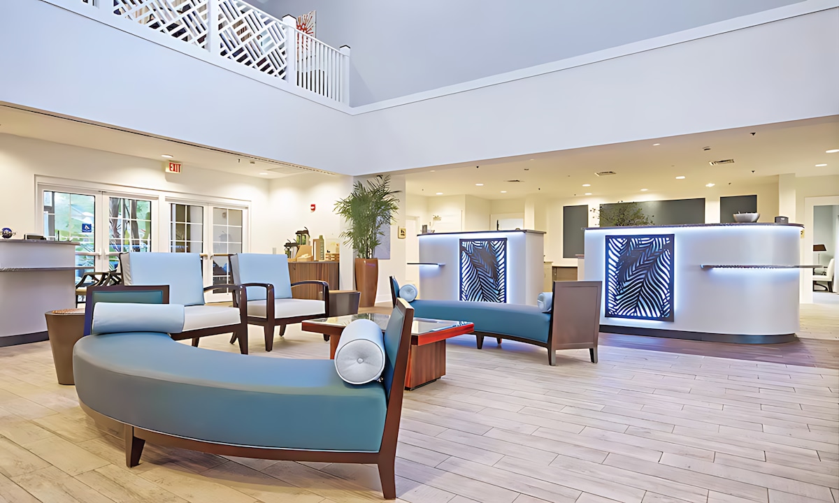 The lobby area features modern seating arrangements, including curved sofas and lounge chairs designed for comfort. A reception counter illuminated with soft lighting is prominently displayed. Large windows allow natural light to brighten the space, creating an open and welcoming environment.