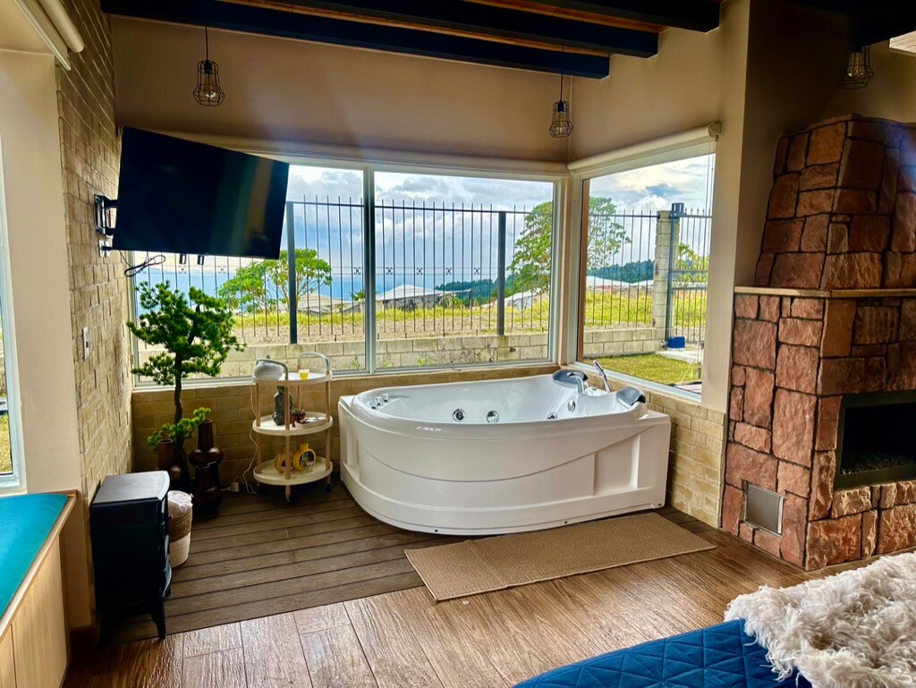 A spacious area featuring a large, modern whirlpool tub positioned near floor-to-ceiling windows, allowing for views of the surrounding nature. A stone fireplace complements the space, while a cozy seating area is present nearby, creating an inviting environment.