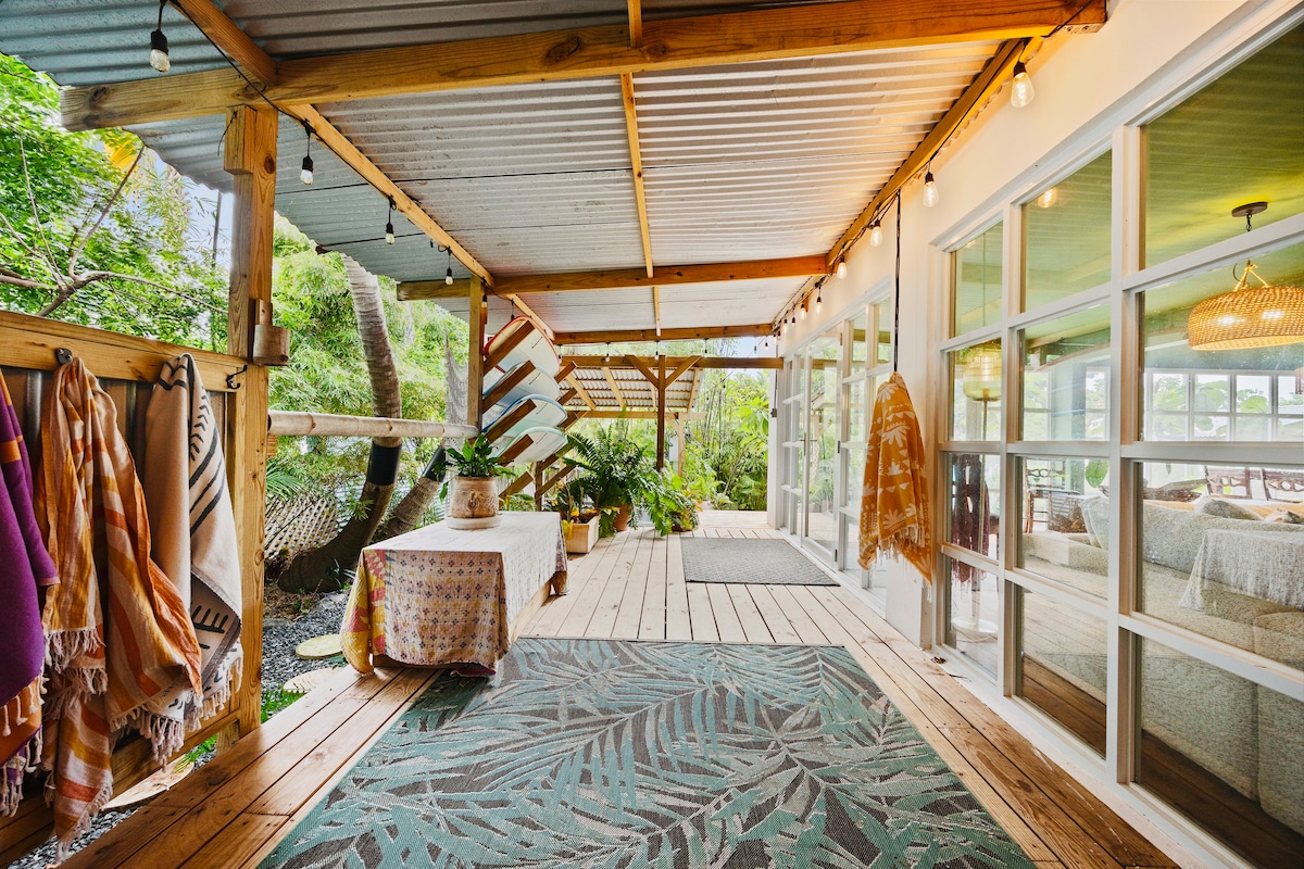 A spacious porch features wooden flooring and a corrugated metal roof, adorned with soft lighting. Potted plants and decorative textiles create a welcoming space that connects to the outdoors, with large windows providing views of the surrounding greenery.