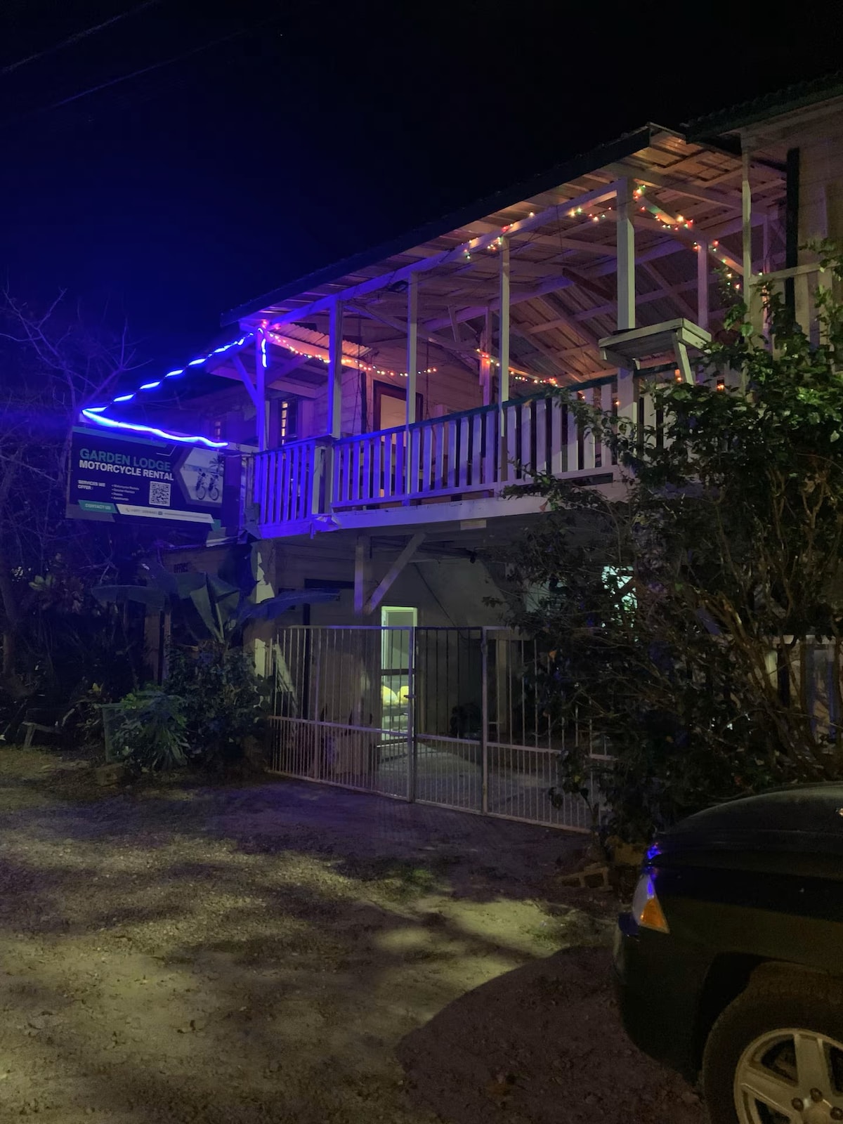 A two-story building is seen at night, with string lights illuminating the exterior. The upper balcony is adorned with colorful lights, enhancing the space. Lush greenery surrounds the entrance, which features a gated pathway leading to the front door.