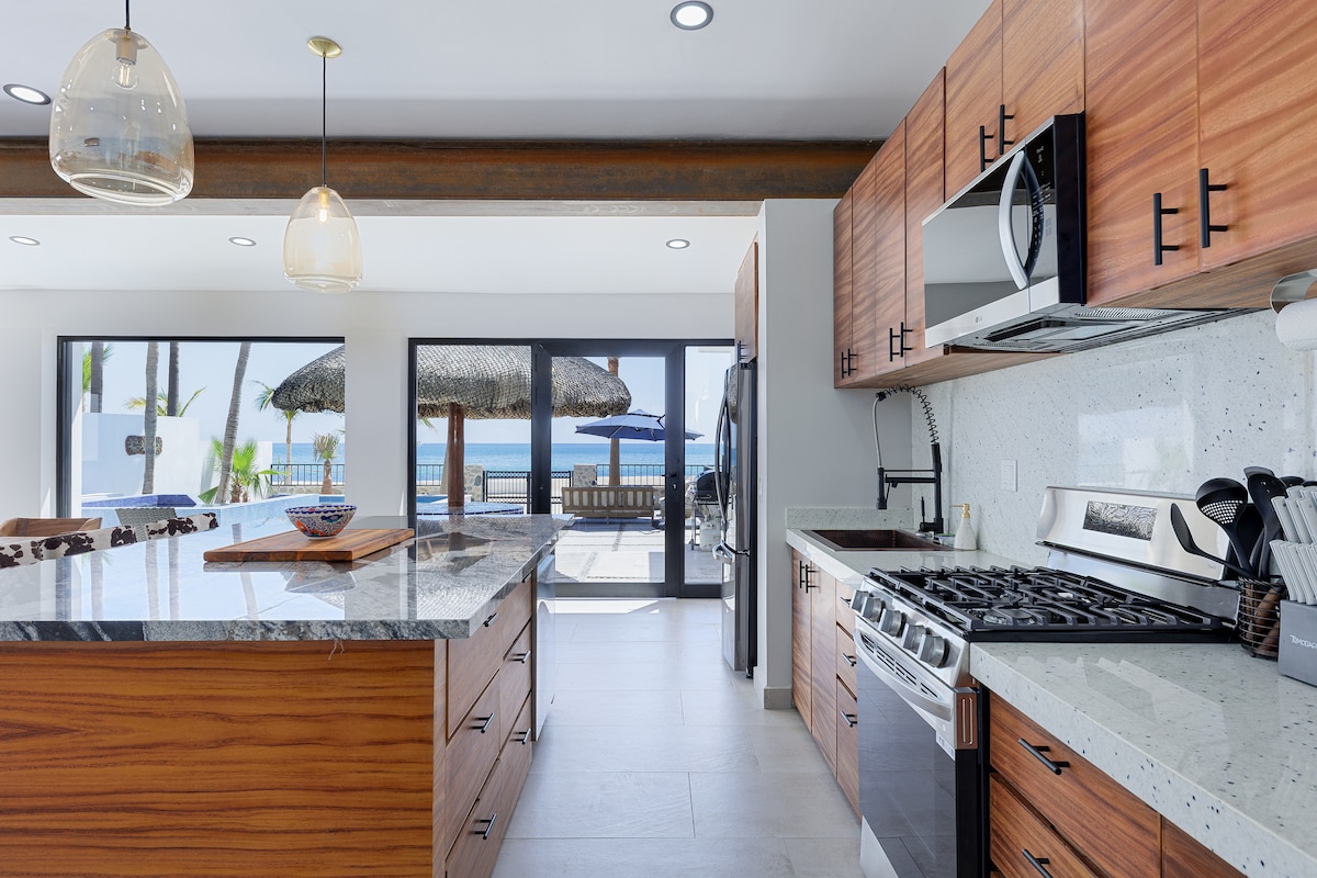 A modern kitchen features sleek wood cabinetry and a spacious island with a wooden serving board. Stainless steel appliances, including a gas stove and microwave, are visible. Large sliding glass doors provide a view of the outdoor space and beach beyond, allowing natural light to fill the room.