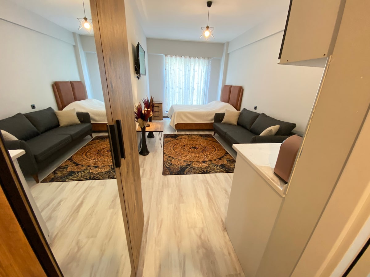 A well-lit room features two spacious beds with minimalistic headboards, flanked by dark sofas and accent tables. A large window is adorned with sheer curtains, allowing natural light to brighten the space. Decorative rugs add warmth to the room's neutral tones.