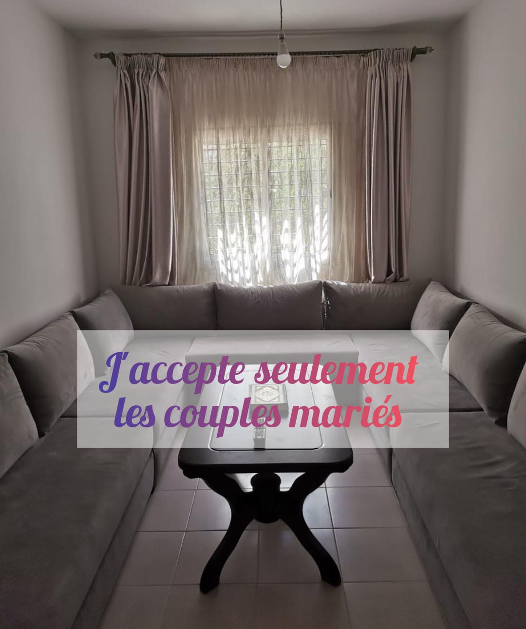 A spacious living area is showcased, featuring a large, U-shaped sofa in neutral tones, arranged around a small, dark wood table. Natural light filters through sheer curtains at the window, illuminating the room's relaxed atmosphere.