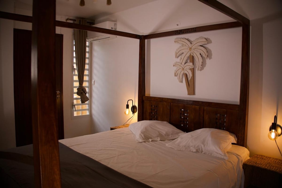 A spacious bedroom features a four-poster bed with a neutral-toned bedding set. A wooden wall decoration of palm trees adds interest. Soft lighting is provided by wall sconces on either side of the bed, while natural light enters through a partially open window with vertical slats.