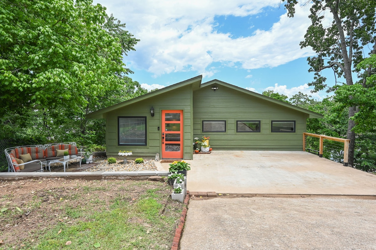 The exterior of the lake house is shown, featuring a green façade complemented by large windows and a distinctive orange door. A cozy seating area with colorful cushions is situated on the porch, surrounded by greenery and decorative potted plants.