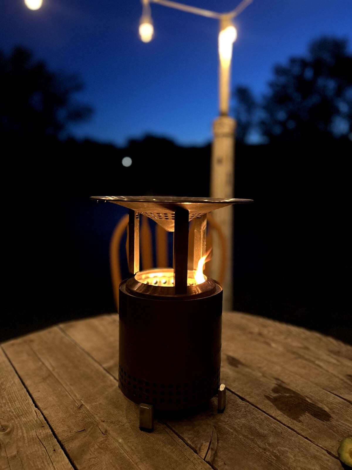 A tabletop fire feature stands on a weathered wooden surface, casting a warm glow in the evening. Soft light from nearby string lights enhances the serene atmosphere, while dark silhouettes of trees are visible in the background under a twilight sky.
